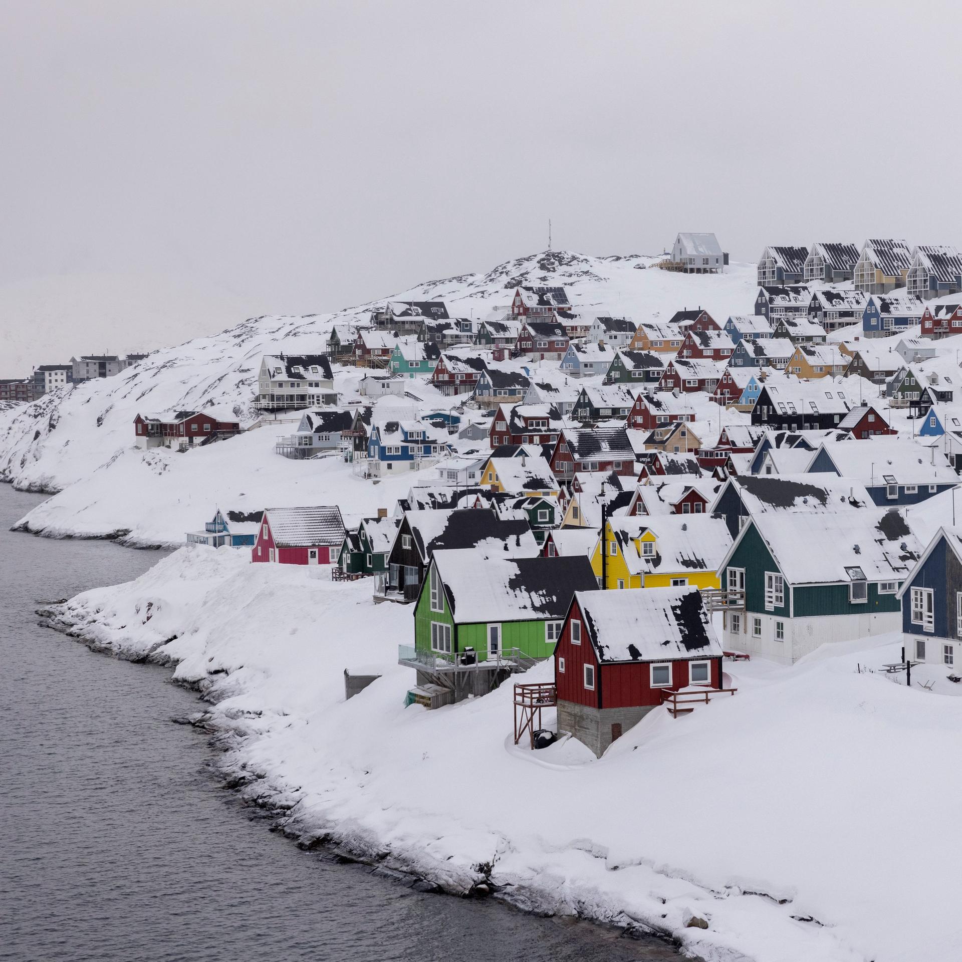 Bunte Holzhäuser stehen dicht gedrängt in der tief verschneiten Hauptstadt Nuuk. Links ist Gewässer zu sehen. 
