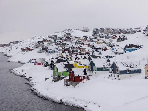 Bunte Holzhäuser stehen dicht gedrängt in der tief verschneiten Hauptstadt Nuuk. Links ist Gewässer zu sehen. 