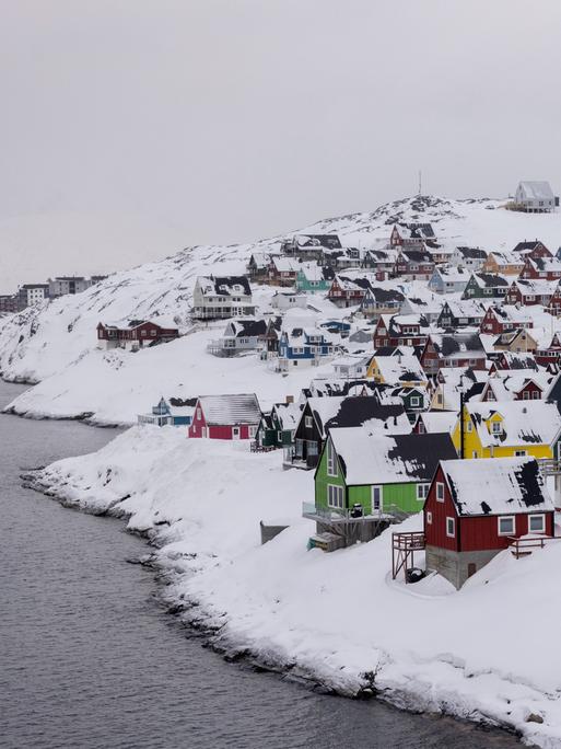 Bunte Holzhäuser stehen dicht gedrängt in der tief verschneiten Hauptstadt Nuuk. Links ist Gewässer zu sehen. 