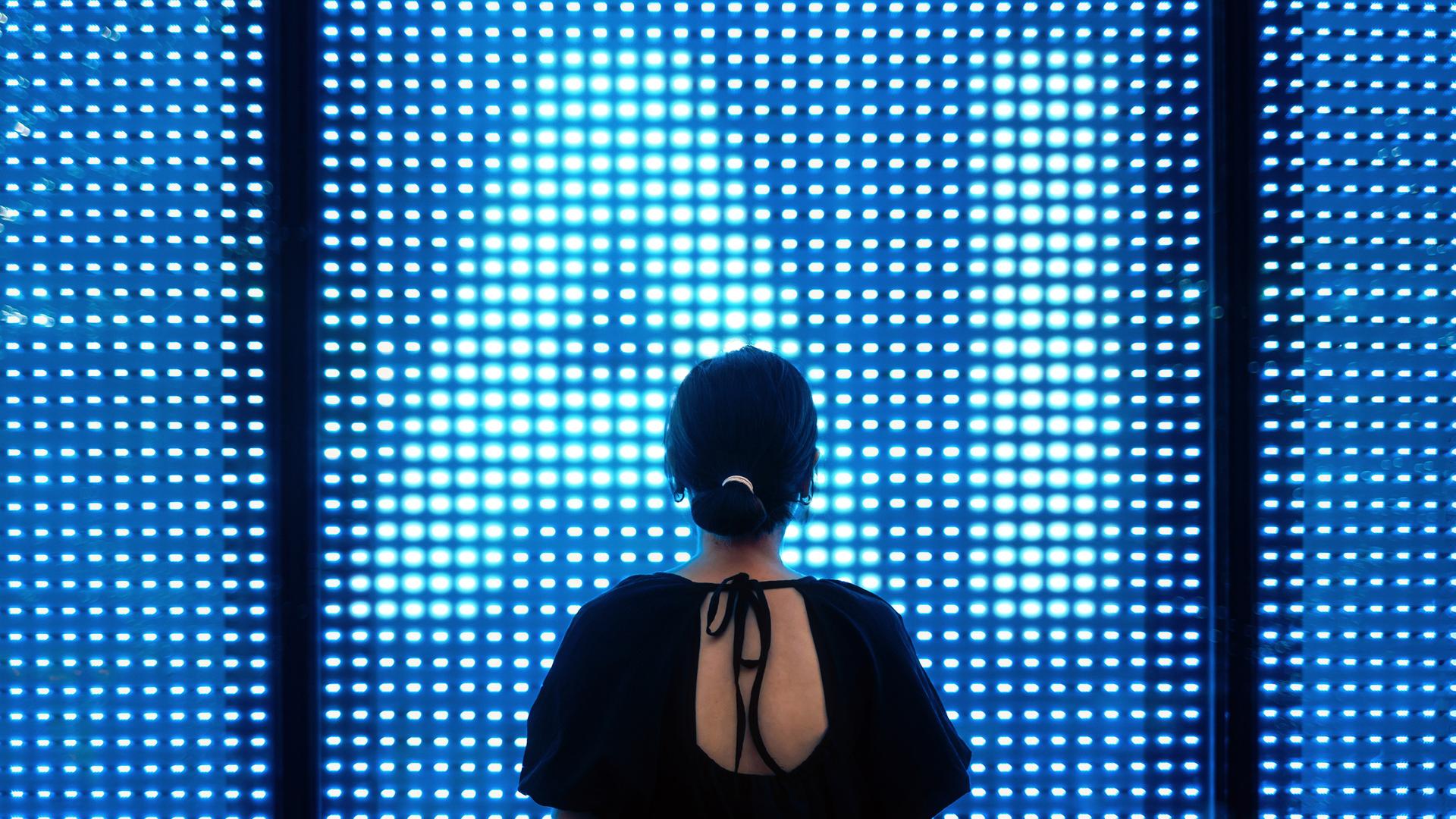 Eine Frau steht dem Betrachter mit dem Rücken zugewandt vor einer erleuchtenden LED Wand, deren Lichtpunkte die Buchstaben "AI" formen. Eine Frau steht dem Betrachter mit dem Rücken zugewandt vor einer erleuchtenden LED Wand, deren Lichtpunkte die Buchstaben "AI" formen.