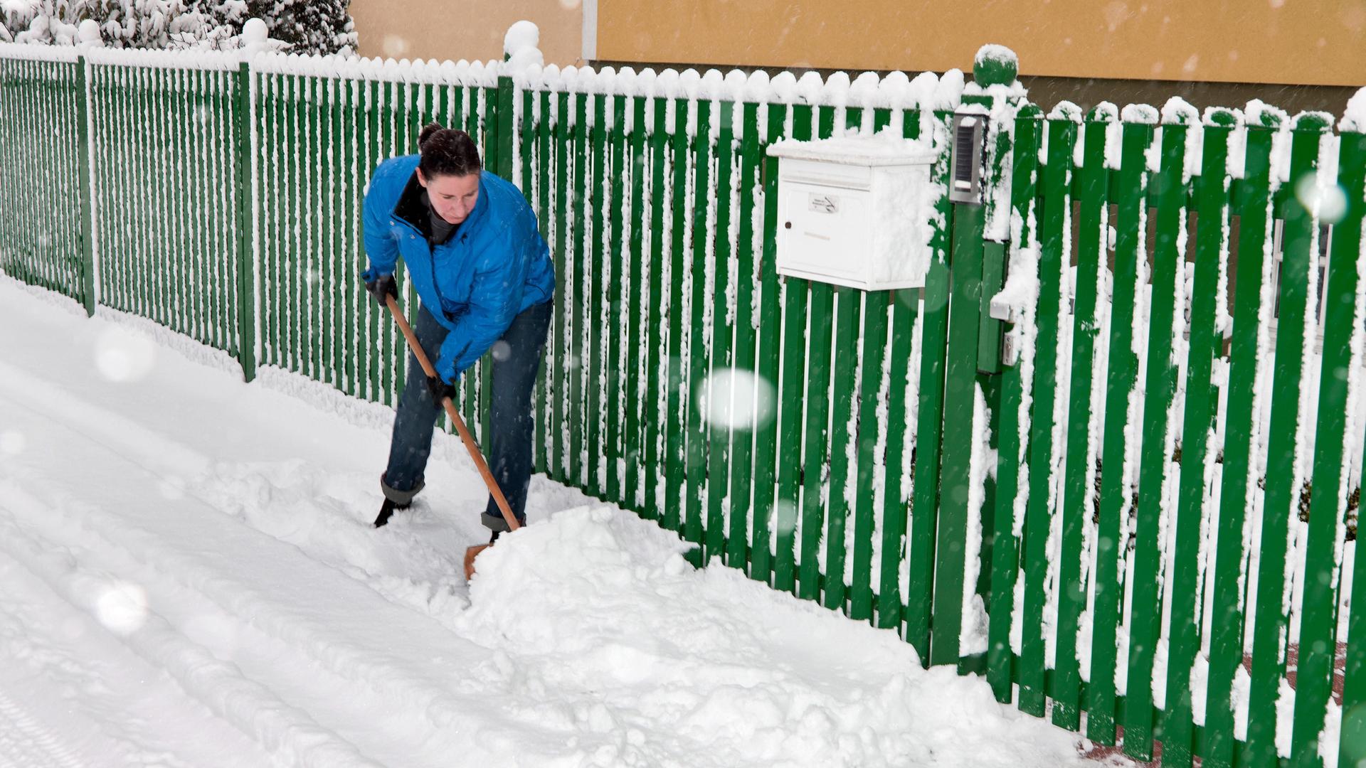 Eine Frau mit blauer Arbeitsjacke schippt Schnee vor einem grünen Zaun