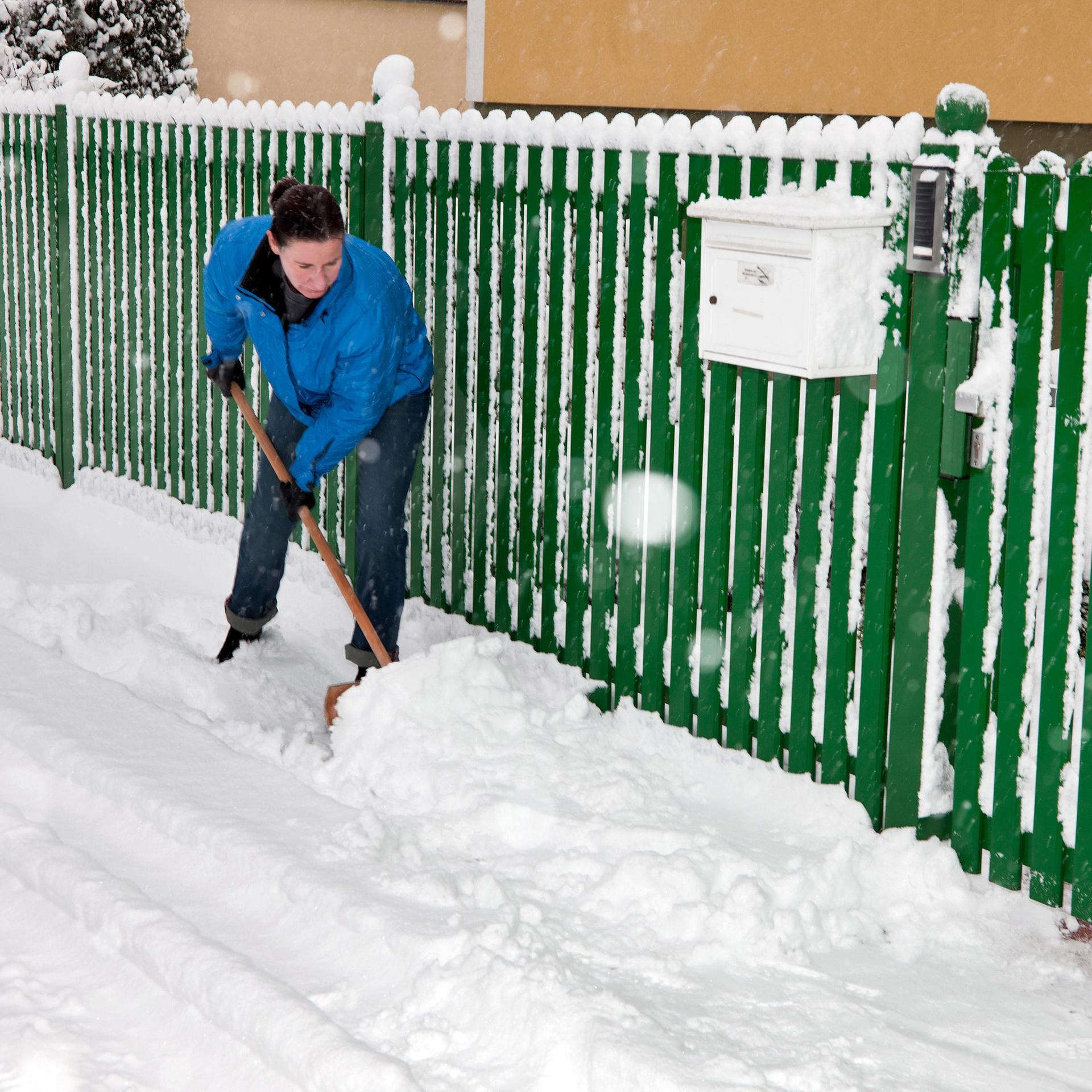 Eine Frau mit blauer Arbeitsjacke schippt Schnee vor einem grünen Zaun