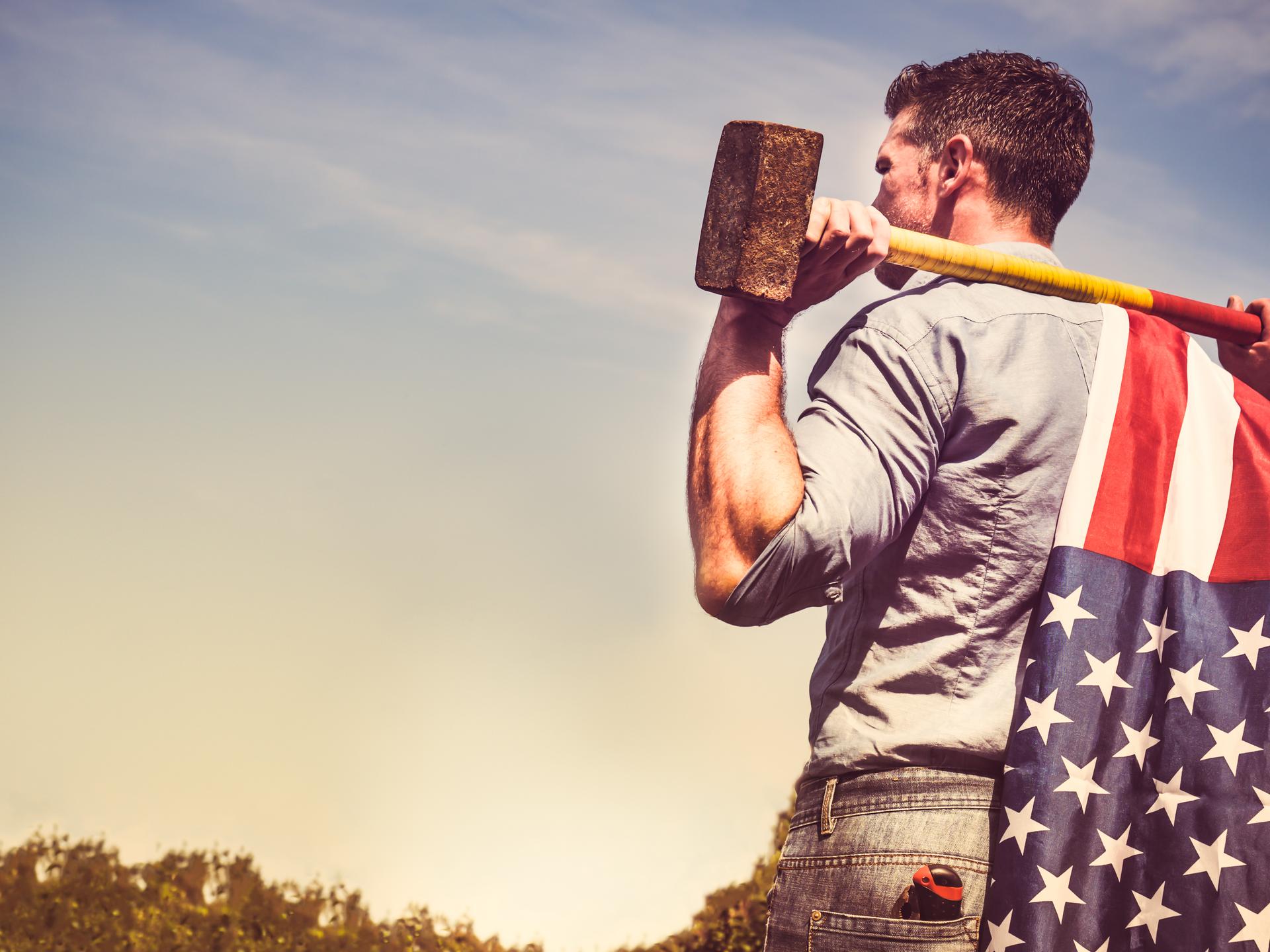 Ein Mann in Hemd und Jeans steht mit einem Vorschlaghammer über die Schultern gelegt. Er guckt in Richtung des Horizonts. Am Stiel des Vorschlaghammers ist eine amerikanische Flagge befestigt.