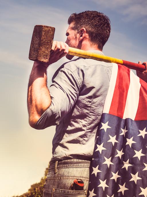 Ein Mann in Hemd und Jeans steht mit einem Vorschlaghammer über die Schultern gelegt. Er guckt in Richtung des Horizonts. Am Stiel des Vorschlaghammers ist eine amerikanische Flagge befestigt.