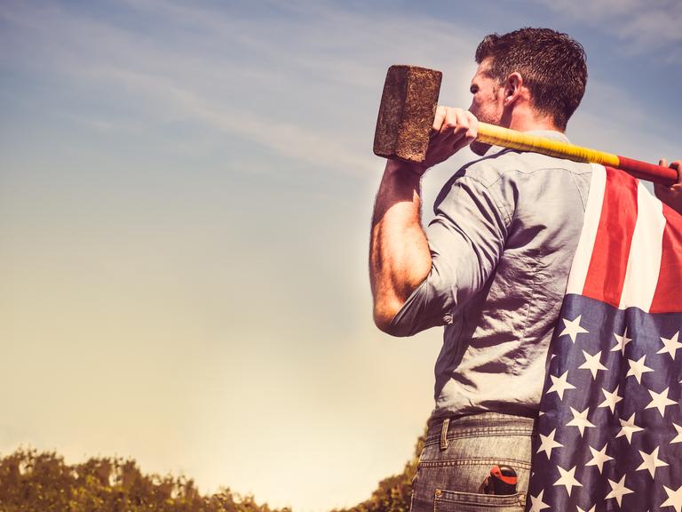 Ein Mann in Hemd und Jeans steht mit einem Vorschlaghammer über die Schultern gelegt. Er guckt in Richtung des Horizonts. Am Stiel des Vorschlaghammers ist eine amerikanische Flagge befestigt.