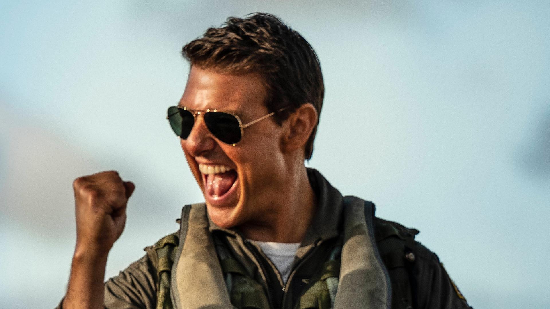 Tom Cruise in der Rolle des Capt. Pete Maverick Mitchell im Film "Top Gun: Maverick" von Joseph Kosinski. Er trägt einen Pilotenanzug und eine dunkle Sonenbrille und ballt die Faust jubelnd.