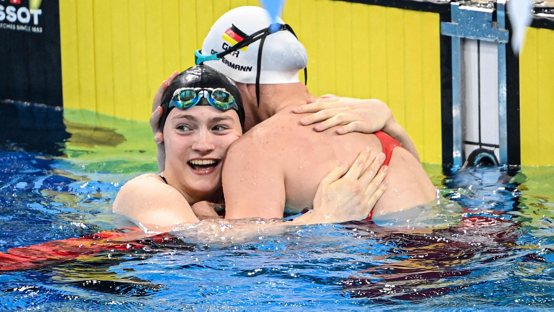 Zwei Frauen mit Bade-Kappen umarmen sich in einem Schwimm-Becken. 