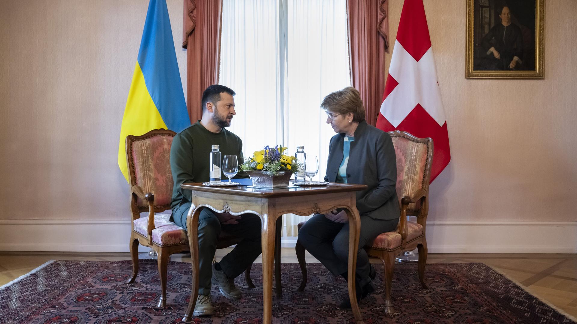 Die Bundespräsidentin der Schweiz, Viola Amherd sitzt mit dem ukrainischen Präsidenten Wolodymyr Selenskyj an einem Tisch und unterhält sich. Im Hintergrund zu sehen sind die Landesflaggen.