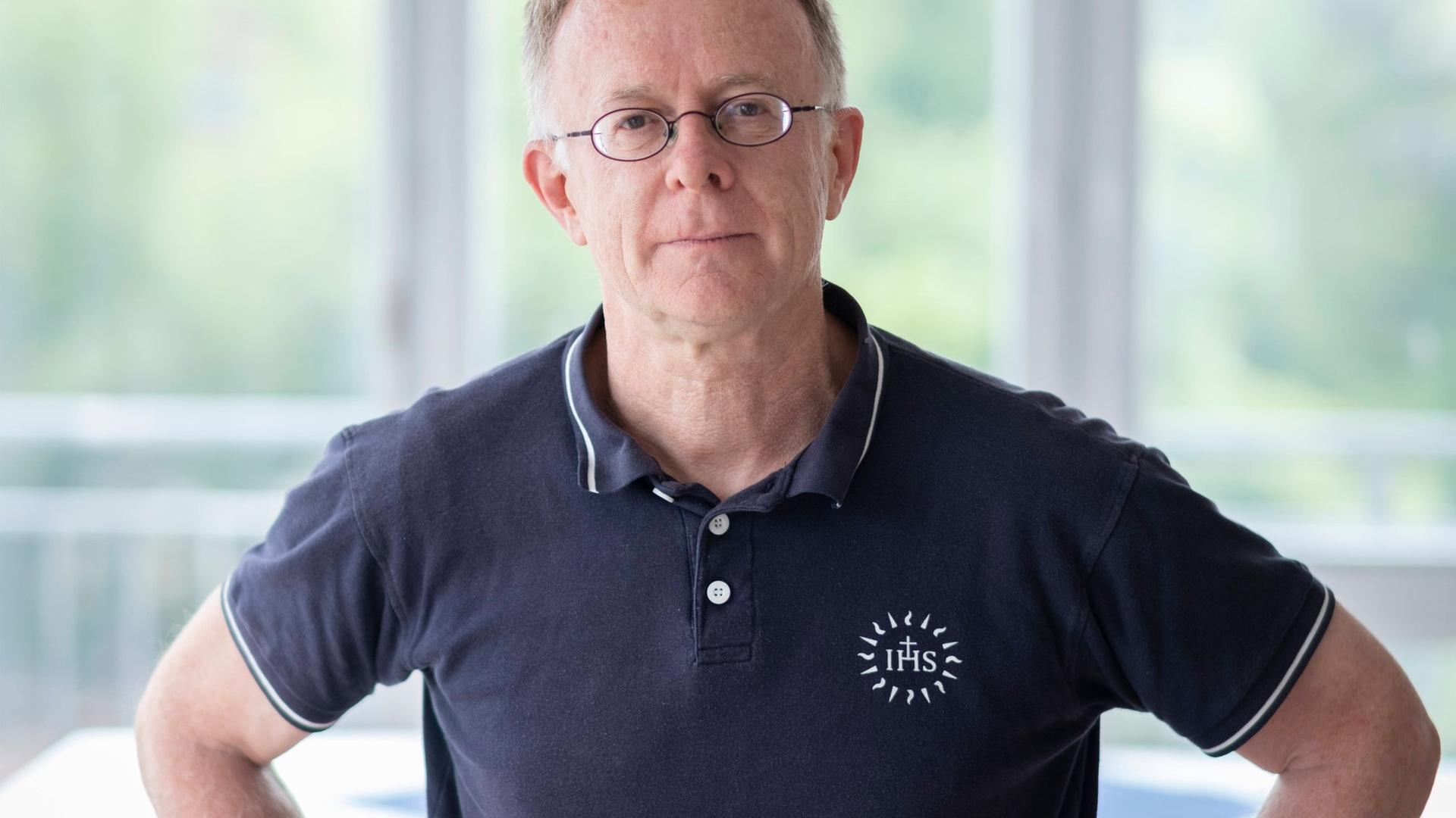Der Jesuitenpater Jörg Alt steht die Arme in den Hüften, in einem hellen Raum mit großen Fenstern. Er trägt ein dunkelblaues Poloshirt und eine Brille und schaut in die Kamera. Der Jesuitenpater Jörg Alt steht die Arme in den Hüften, in einem hellen Raum mit großen Fenstern. Er trägt ein dunkelblaues Poloshirt und eine Brille und schaut in die Kamera.