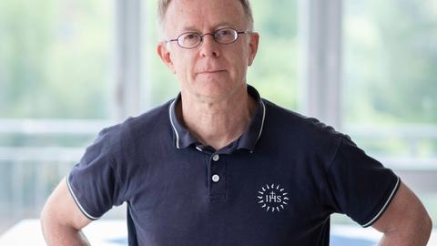 Der Jesuitenpater Jörg Alt steht die Arme in den Hüften, in einem hellen Raum mit großen Fenstern. Er trägt ein dunkelblaues Poloshirt und eine Brille und schaut in die Kamera.