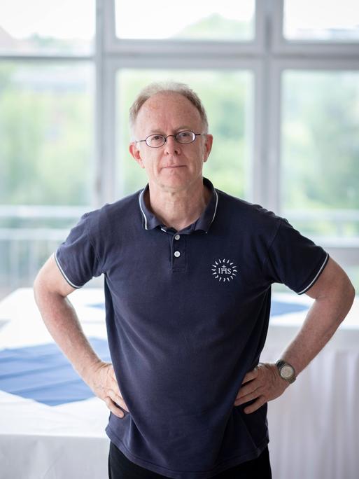 Der Jesuitenpater Jörg Alt steht die Arme in den Hüften, in einem hellen Raum mit großen Fenstern. Er trägt ein dunkelblaues Poloshirt und eine Brille und schaut in die Kamera.