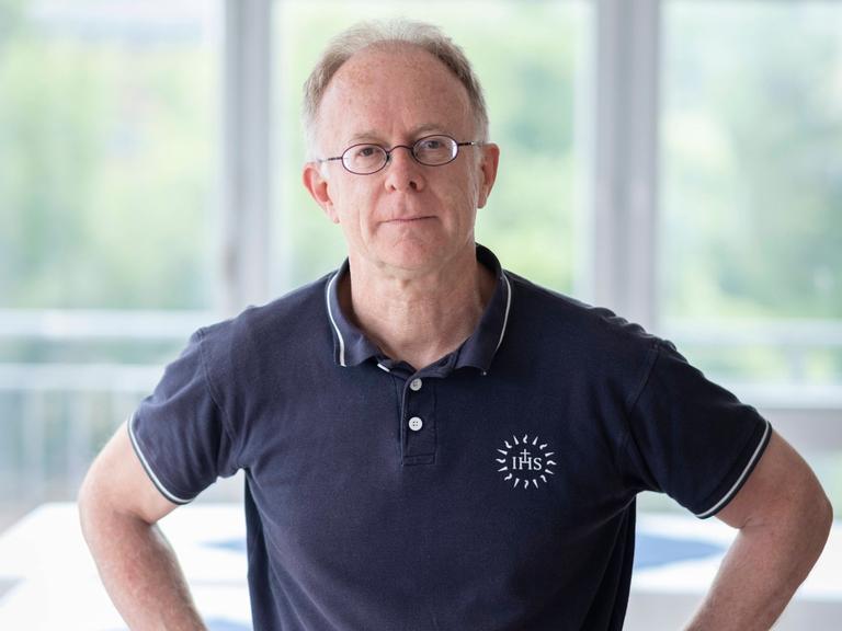 Der Jesuitenpater Jörg Alt steht die Arme in den Hüften, in einem hellen Raum mit großen Fenstern. Er trägt ein dunkelblaues Poloshirt und eine Brille und schaut in die Kamera.
