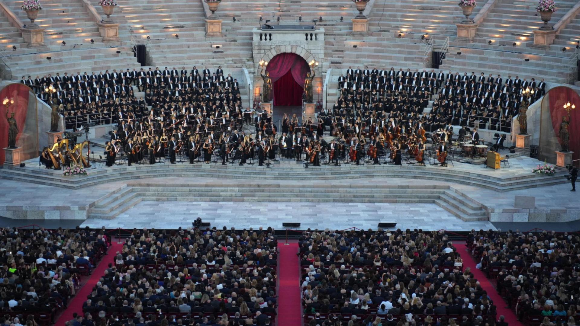 Zu sehen ist eine Bühne in einer alten, italienischen Arena. Auf der Bühne stehen Dutzende Menschen in schwarzer, eleganter Kleidung. Davor sieht man einen Teil des Publikums.