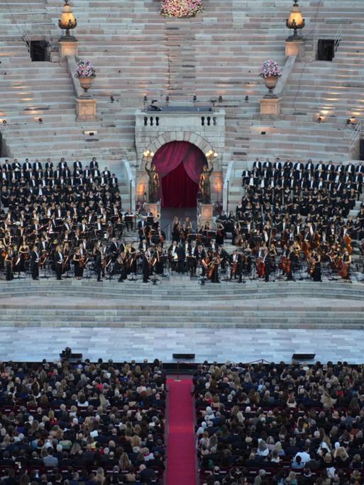 Zu sehen ist eine Bühne in einer alten, italienischen Arena. Auf der Bühne stehen Dutzende Menschen in schwarzer, eleganter Kleidung. Davor sieht man einen Teil des Publikums.