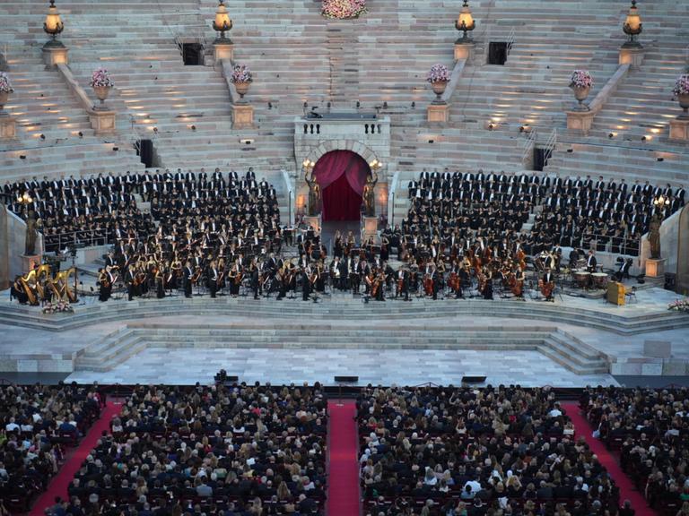 Zu sehen ist eine Bühne in einer alten, italienischen Arena. Auf der Bühne stehen Dutzende Menschen in schwarzer, eleganter Kleidung. Davor sieht man einen Teil des Publikums.