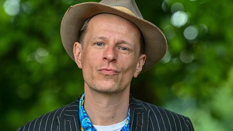 Der Schauspieler Alexander Scheer schaut in die Kamera, im Hintergrund leuchtet das Sommergrün eines Parks.