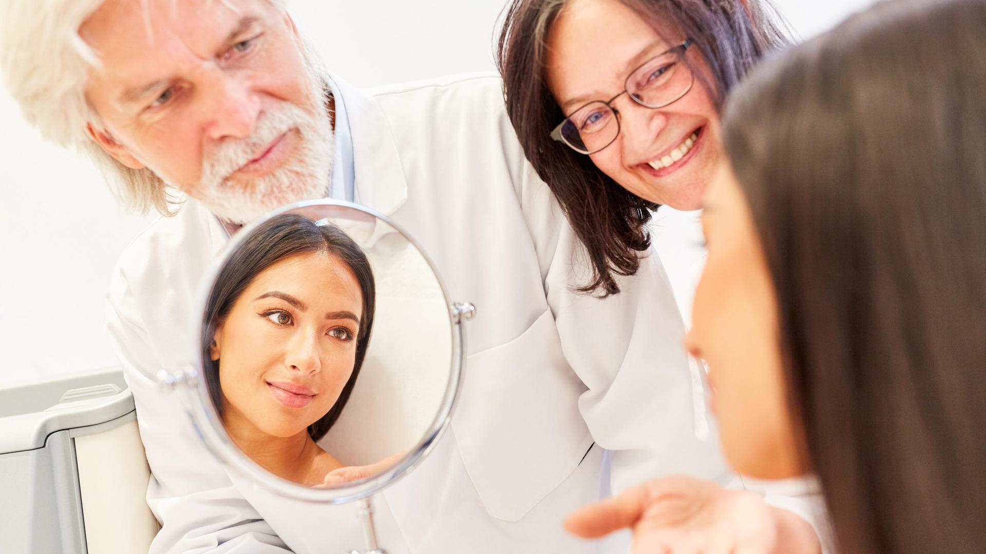 Ãrzteteam der Schönheitsklinik mit Spiegel im Beratungsgespräch mit einer Patientin || Modellfreigabe vorhanden