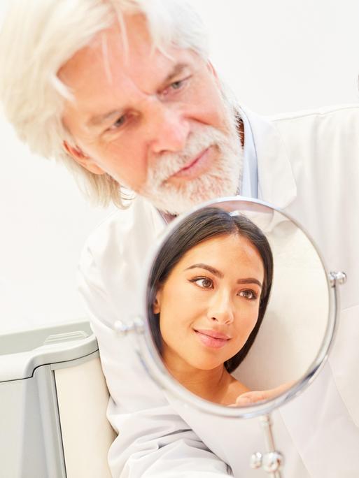Ãrzteteam der Schönheitsklinik mit Spiegel im Beratungsgespräch mit einer Patientin || Modellfreigabe vorhanden