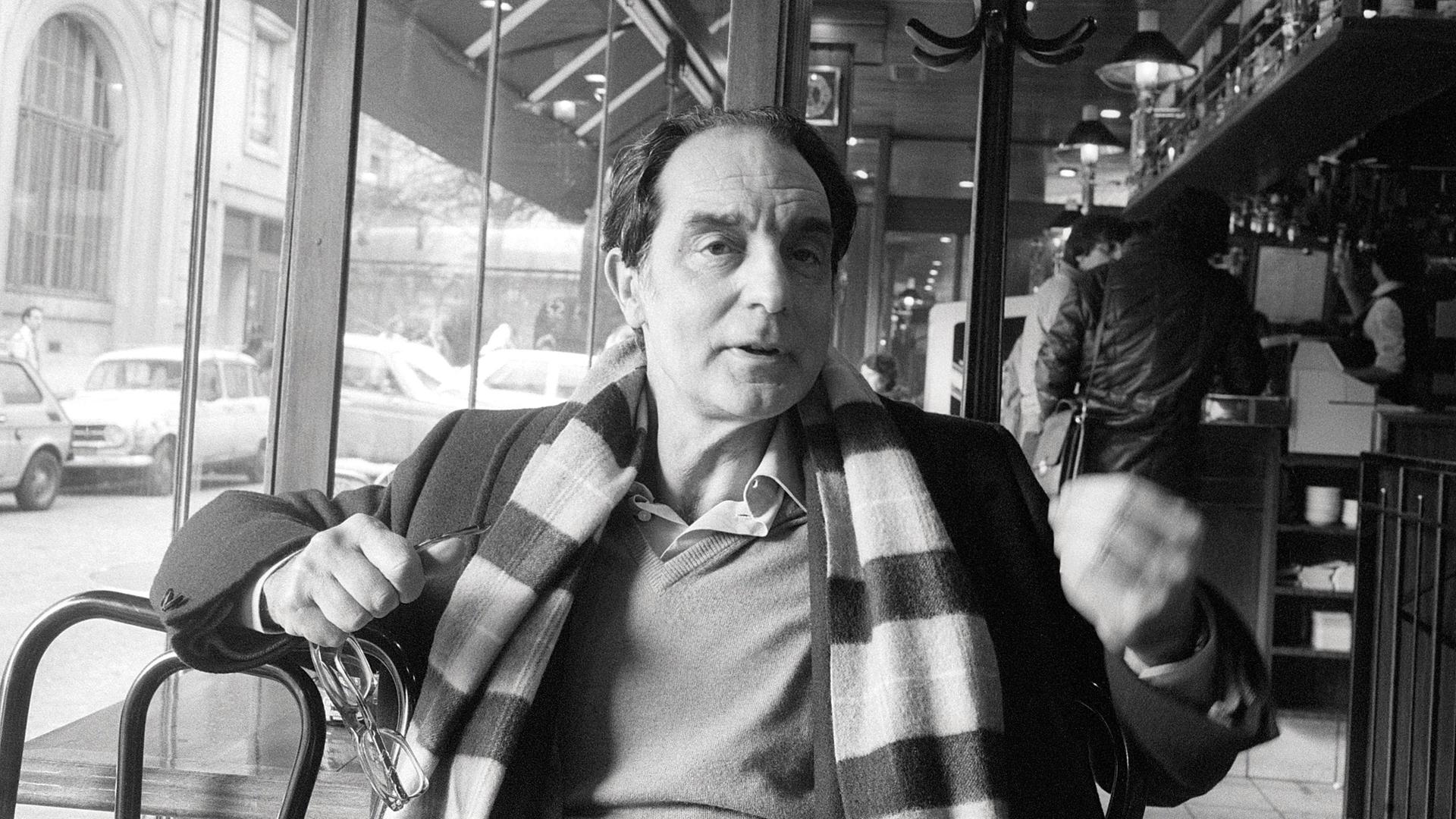 Der Autor Italo Calvino sitzt in einem Café, und gestikuliert. Er trägt einen  breit geringelten Schal.