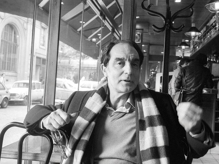 Der Autor Italo Calvino sitzt in einem Café, und gestikuliert. Er trägt einen  breit geringelten Schal.