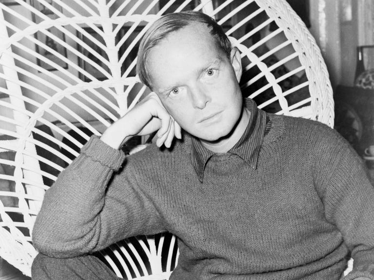 Truman Capote sitzt, seinen Kopf auf die Hand gestützt, auf einem Korbstuhl und schaut in die Kamera