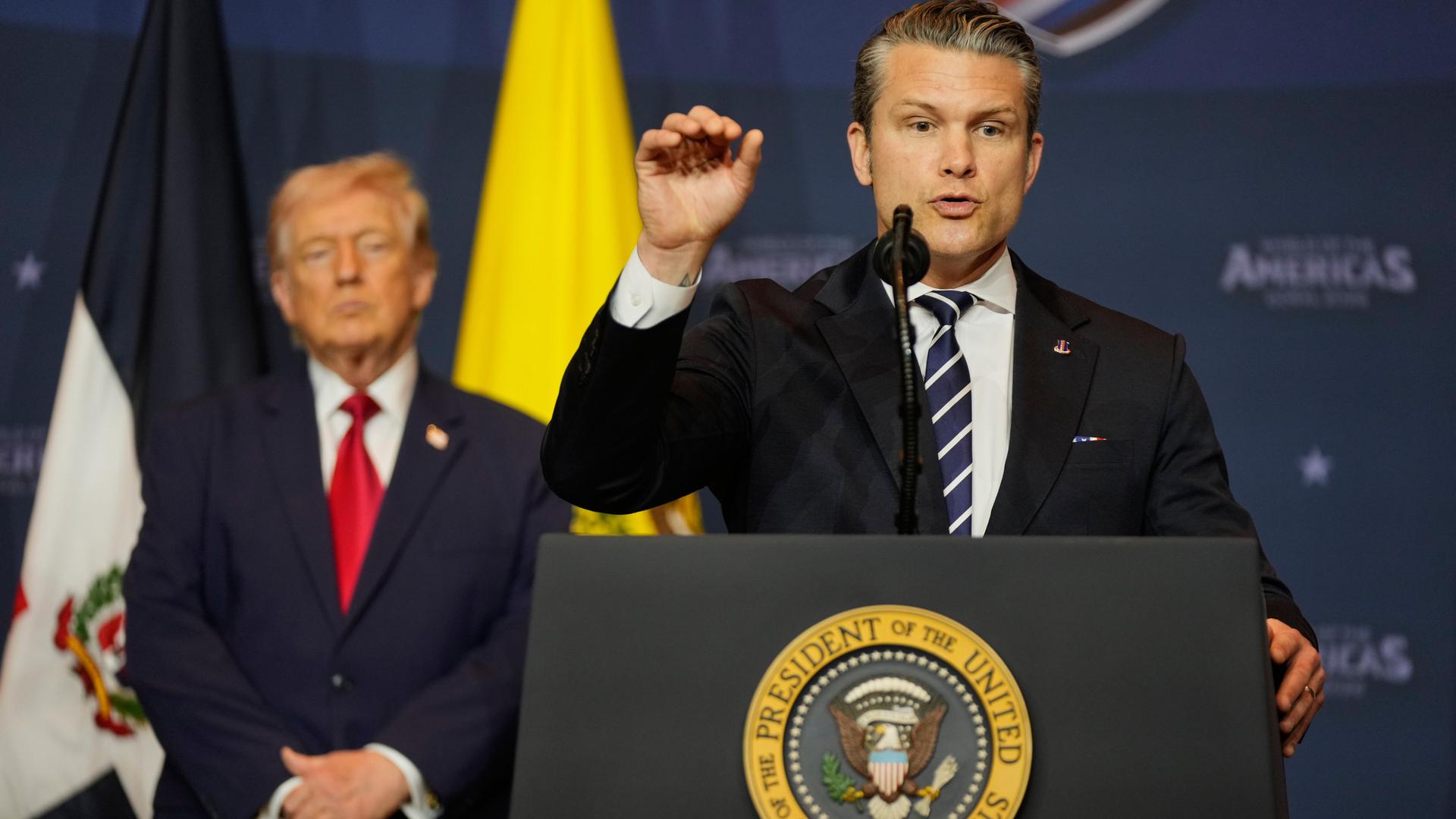 US-Verteidigungsminister Pete Hegseth spricht an einem Rednerpult. Im Hintergrund ist Donald Trump und die US-Flagge zu sehen.