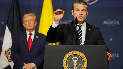 US-Verteidigungsminister Pete Hegseth spricht an einem Rednerpult. Im Hintergrund ist Donald Trump und die US-Flagge zu sehen.