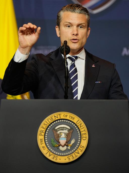 US-Verteidigungsminister Pete Hegseth spricht an einem Rednerpult. Im Hintergrund ist Donald Trump und die US-Flagge zu sehen.