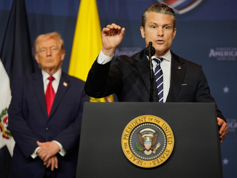 US-Verteidigungsminister Pete Hegseth spricht an einem Rednerpult. Im Hintergrund ist Donald Trump und die US-Flagge zu sehen.