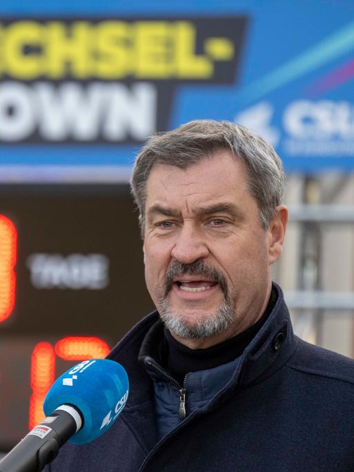 Der CSU-Parteivorsitzende Markus Söder steht im Freien mit geöffnetem Mund vor einem Mikrofon. Im Hintergrund ist ein Digitalbanner mit dem Schriftzug "Politikwechsel Countdown" zu sehen und eine Uhr, die  die Zeit bis zur Bundestagswahl zeigt.