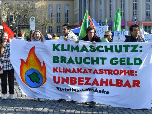 Zwei junge Frauen und drei junge Männer halten ein Transparent auf dem steht: "Klimaschutz braucht Geld. Klimakatastrophe: unbezahlbar. Jetzt mach hin, Anke."