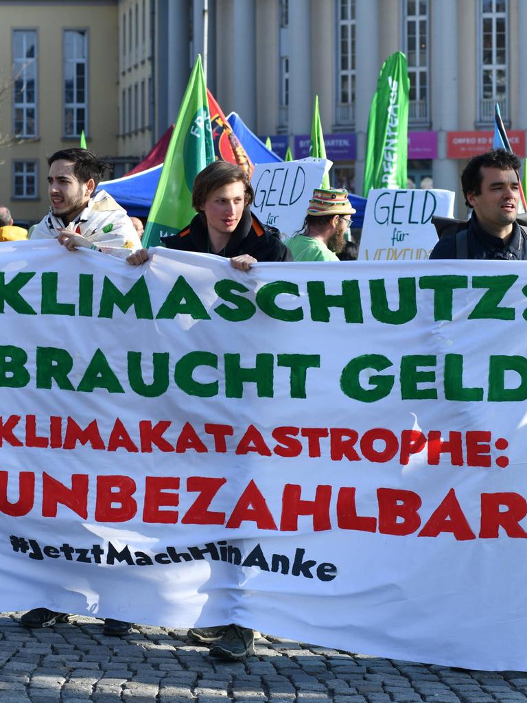 Zwei junge Frauen und drei junge Männer halten ein Transparent auf dem steht: "Klimaschutz braucht Geld. Klimakatastrophe: unbezahlbar. Jetzt mach hin, Anke."