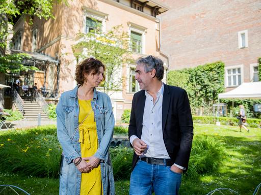 Eva Menasse und Deniz Yücel stehen im Garten des Literaturhauses Berlin und lächeln sich an.