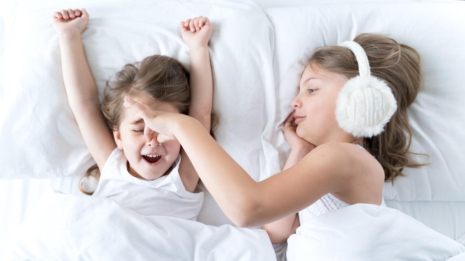 Ein kleines Mädchen liegt im Bett und schnarcht, seine Schwester trägt Kopfhörer und hält ihm die Nase zu