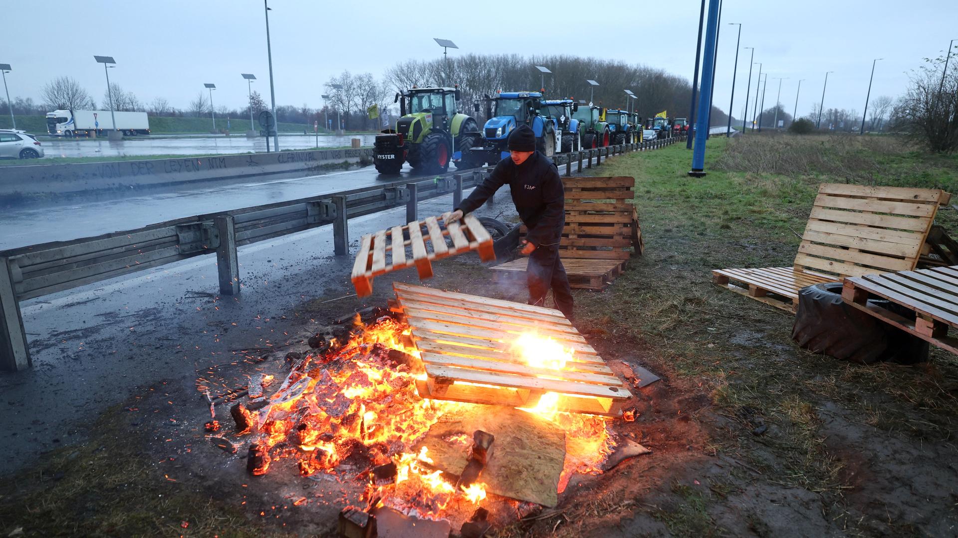 Traktoren blockieren eine Straße. Hinter dem Leitplanke legt ein Mann Holzpaletten auf eine Feuerstelle.