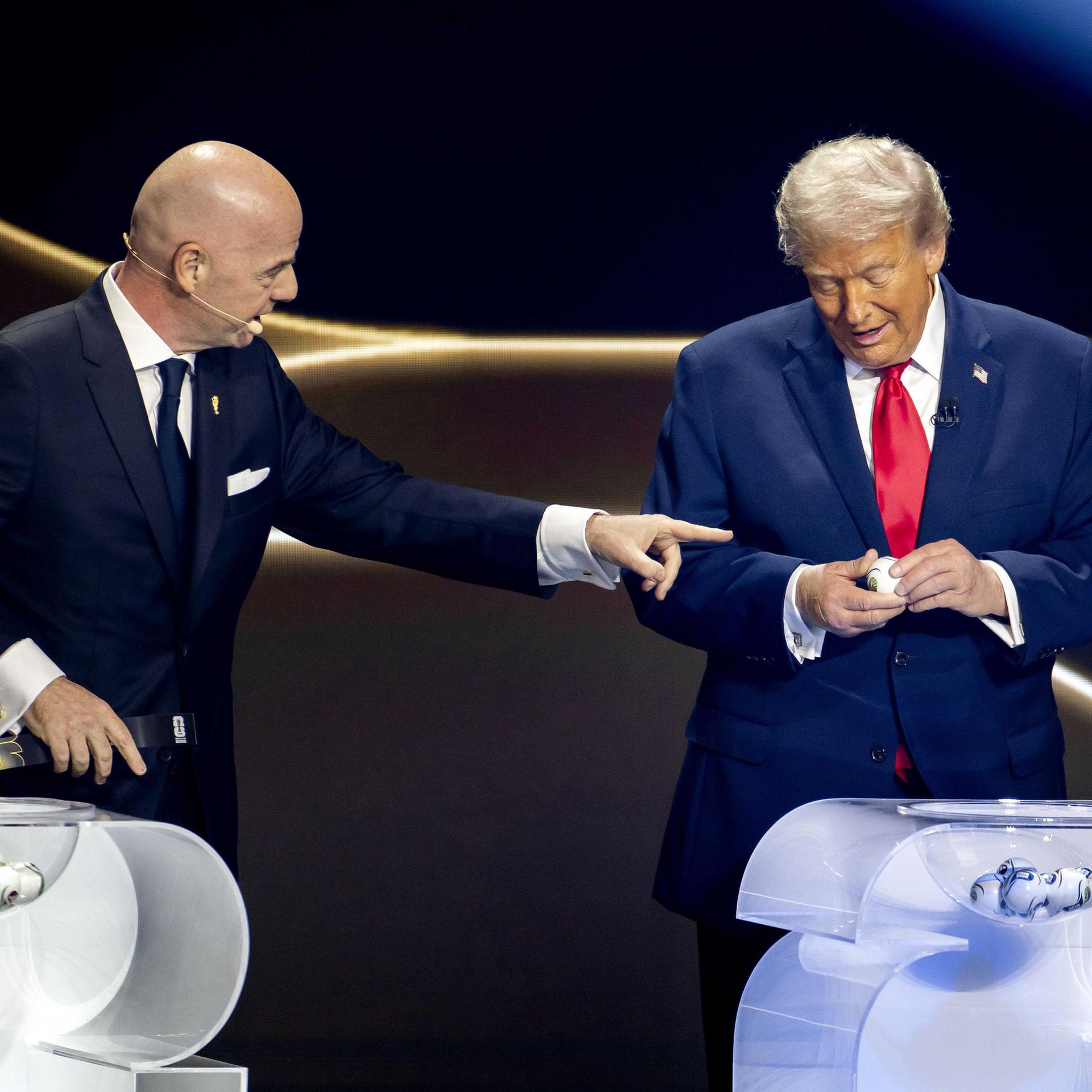 Gianni Infantino und Donald Trump stehen auf einer Bühne vor zwei Lostöpfen. Trump ist dabei, ein Los zu öffnen. Infantino zeigt auf ihn.