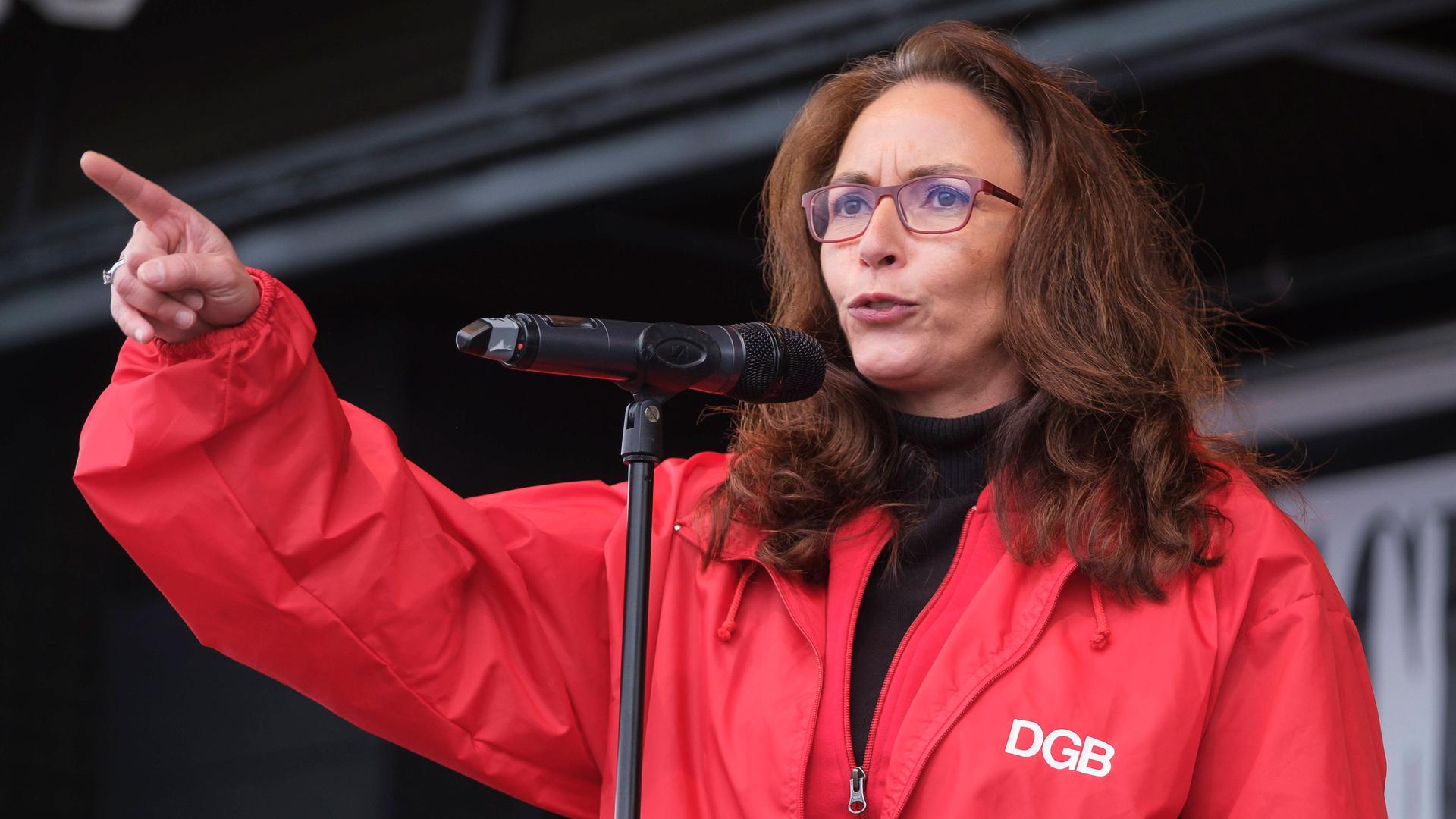 Die Vorsitzende des DGB Yasmin Fahimi: Sie hat eine rote Jacke an mit dem Schriftzug DGB. Fahimi hat lange braune Haare und eine Brille. Sie spricht in ein Mikrofon. Ihr rechter Arm ist erboben, der Zeigefinger ausgetreckt. 