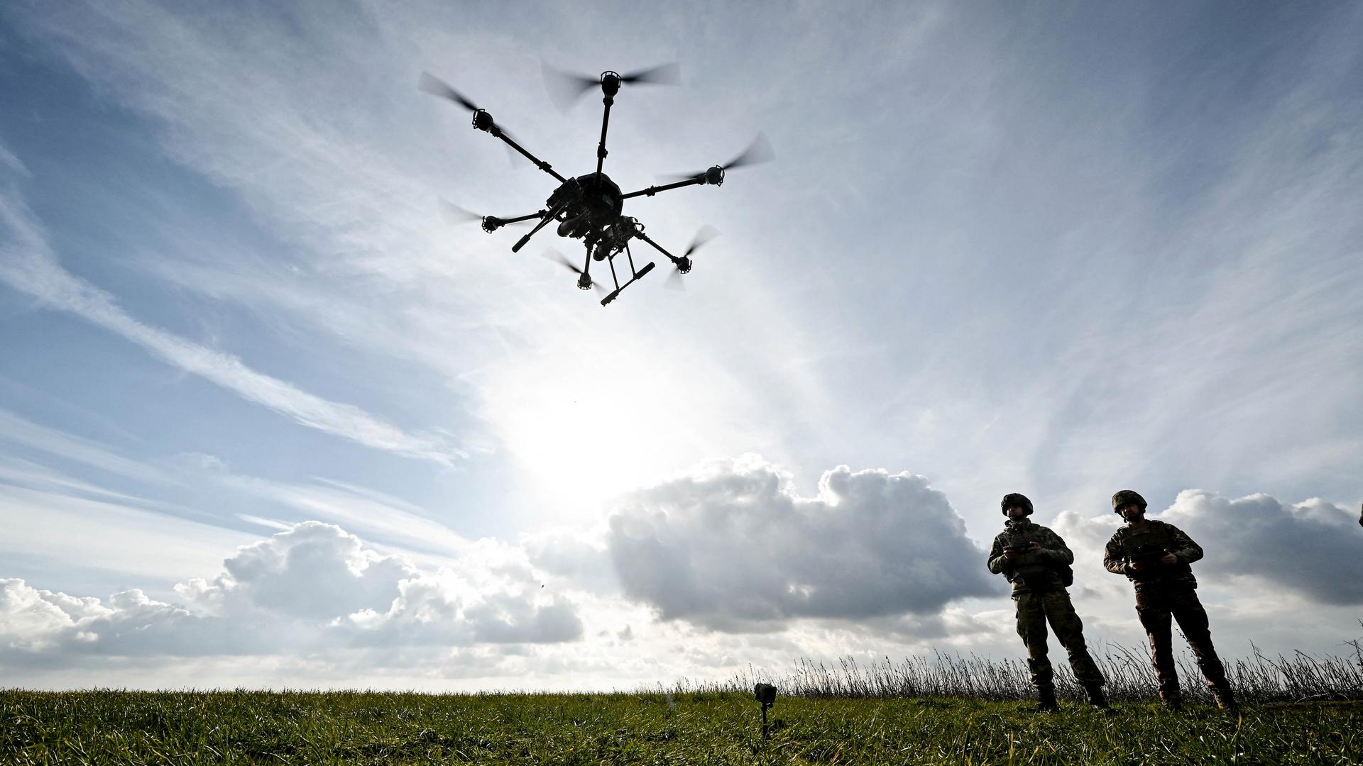 Zwei Soldaten stehen im Gegenlicht unter wolkigem Himmel auf einer Wiese. Im Vordergrund die schwarze Silhouette einer Drohne in der Luft. 