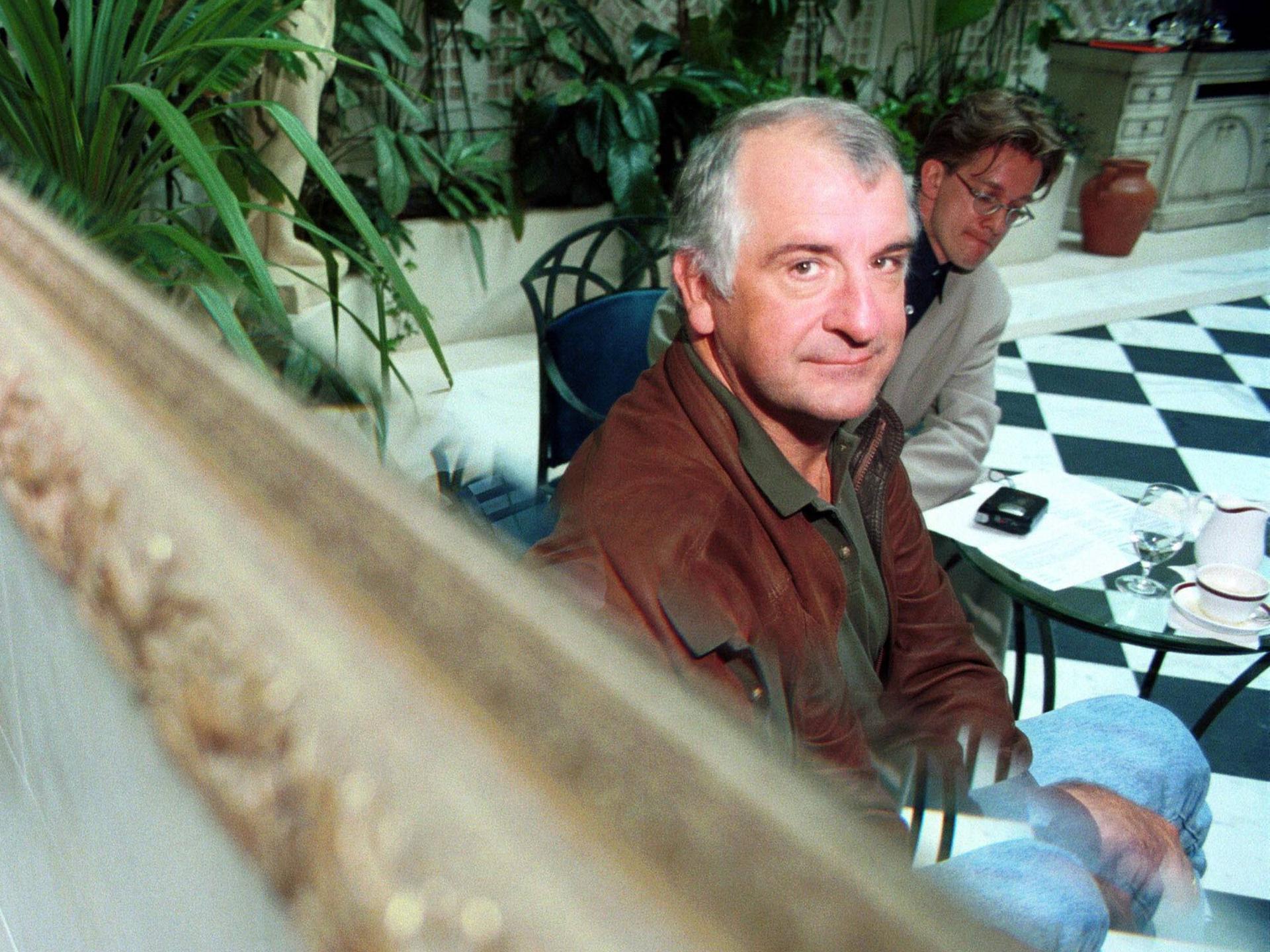 Douglas Adams sitzt in einer edlen Hotellobby und blickt über einen Spiegel in die Kamera