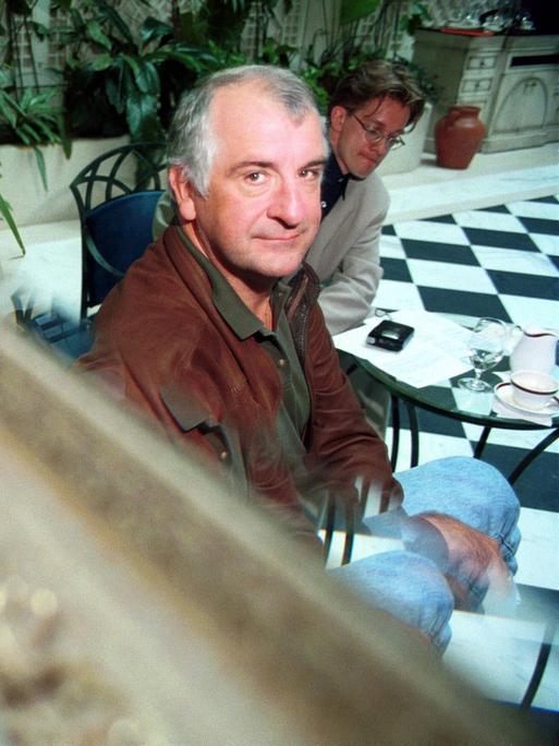 Douglas Adams sitzt in einer edlen Hotellobby und blickt über einen Spiegel in die Kamera