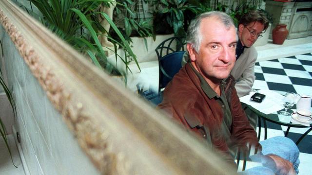Douglas Adams sitzt in einer edlen Hotellobby und blickt über einen Spiegel in die Kamera