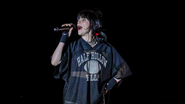 Billie Eilish singt auf einer dunklen Bühne in ein Mikrofon.