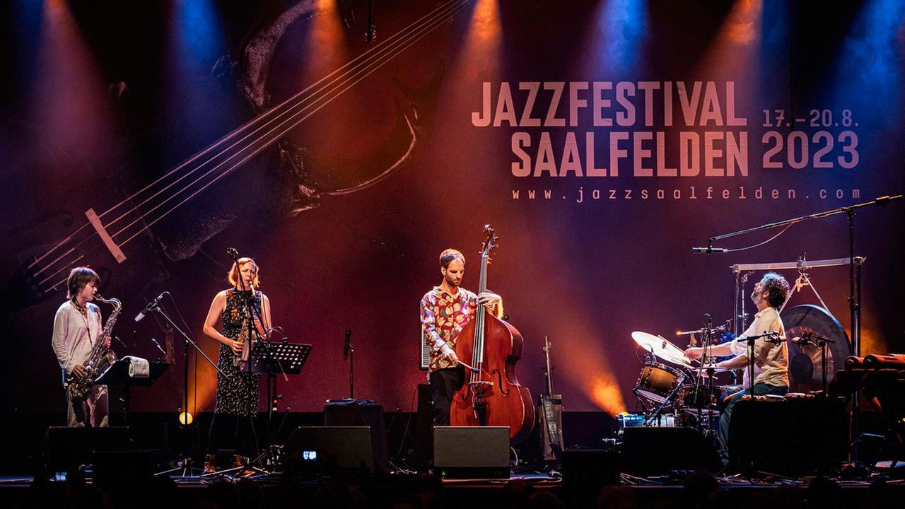 Auf einer großen in buntes Licht gehüllten Bühne spielen zwei Saxofonistinnen, ein Bassist und ganz rechts ein Schlagzeuger unter dem Logo vom Saalfeldener Jazzfestival.