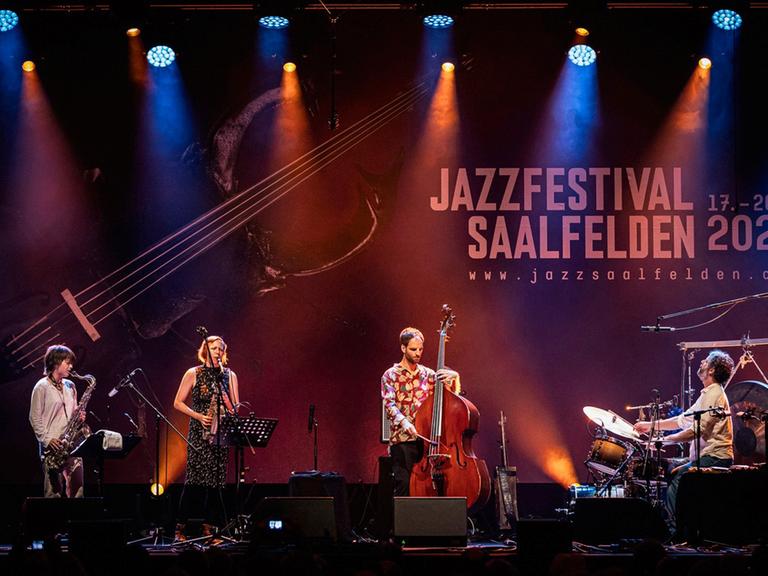 Auf einer großen in buntes Licht gehüllten Bühne spielen zwei Saxofonistinnen, ein Bassist und ganz rechts ein Schlagzeuger unter dem Logo vom Saalfeldener Jazzfestival.