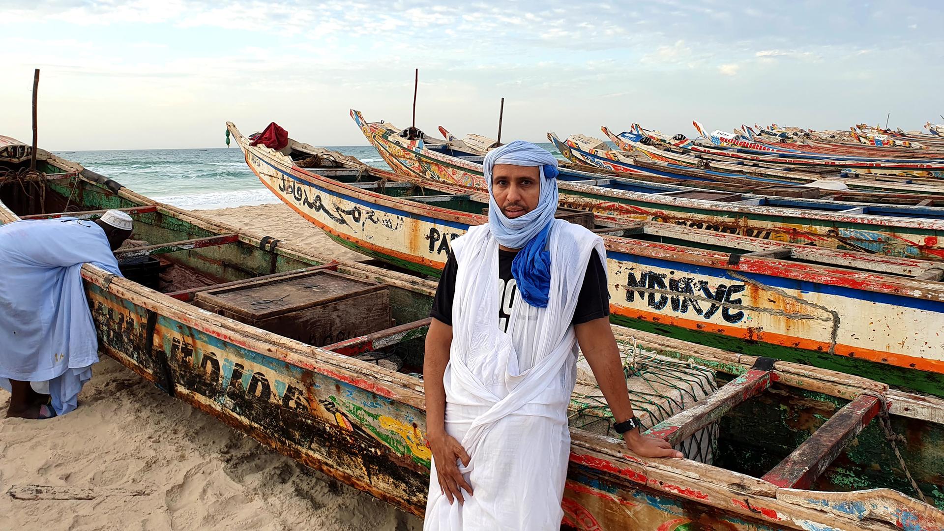 Mohamedou Slahi steht mit Kopftuch bekleidet zwischen bunten Booten am Strand Mohamedou Slahi steht mit Kopftuch bekleidet zwischen bunten Booten am Strand