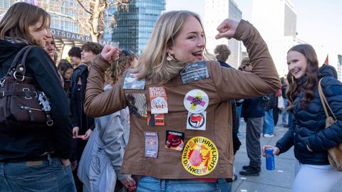 Schülerin hebt bei einer Schülerdemo lachend die Arme und zeigt ihre Jacke mit Aufklebern gegen Wehrpflicht und AfD sowie zum Schulstreik und der Antifa