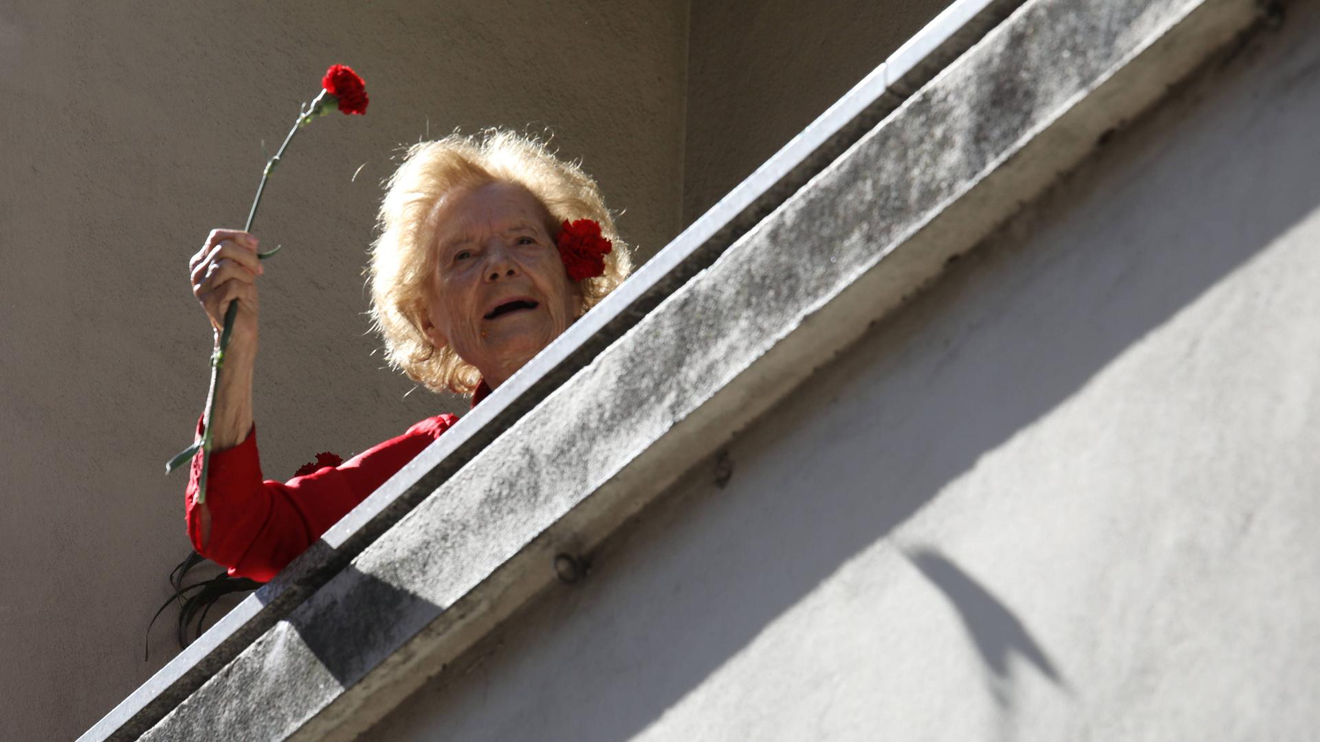 Jährlich wird der Nelkenrevolution in Portugal gedacht. Eine ältere Dame auf dem Balkon schwenkt eine rote Nelke. 