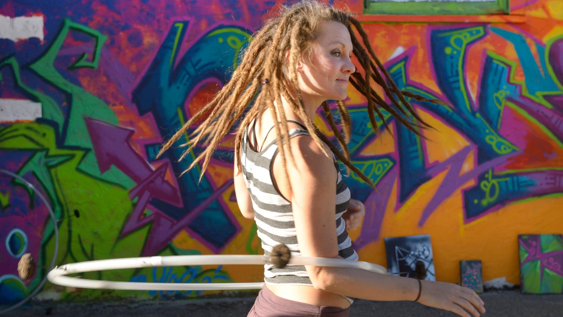 Eine junge Frau trägt Dreadlocks und dreht sich mit ihrem Hula-hoop Reifen  vor einem großen Graffiti.