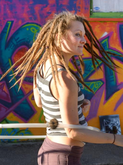 Eine junge Frau trägt Dreadlocks und dreht sich mit ihrem Hula-hoop Reifen  vor einem großen Graffiti.