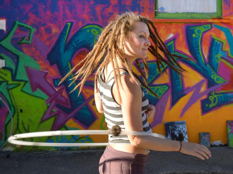 Eine junge Frau trägt Dreadlocks und dreht sich mit ihrem Hula-hoop Reifen  vor einem großen Graffiti.